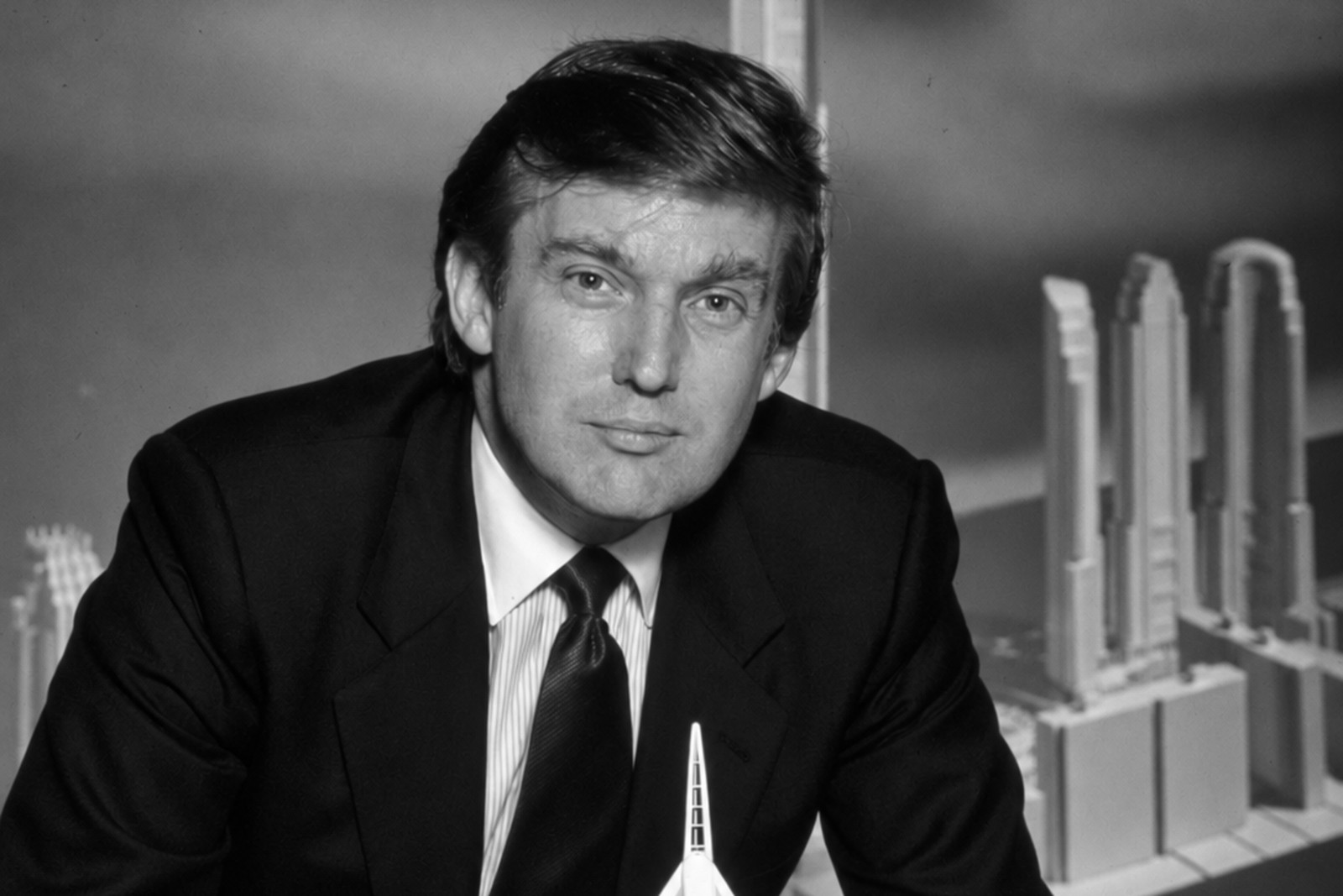 Donald Trump de terno escuro, inclinado sobre maquetes brancas de arranha-céus, com a ponta de uma torre em primeiro plano.