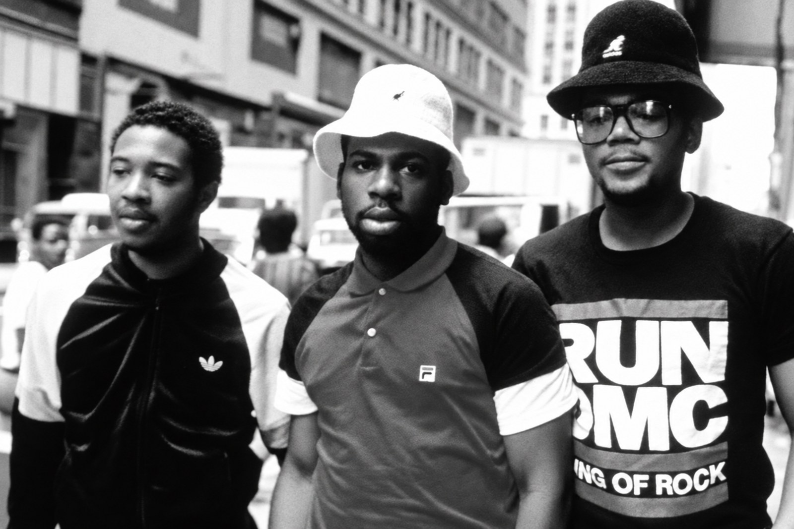 Três homens de chapéu e óculos caminhando por uma rua movimentada de Nova York nos anos 1980, um deles com camiseta estampada do grupo.