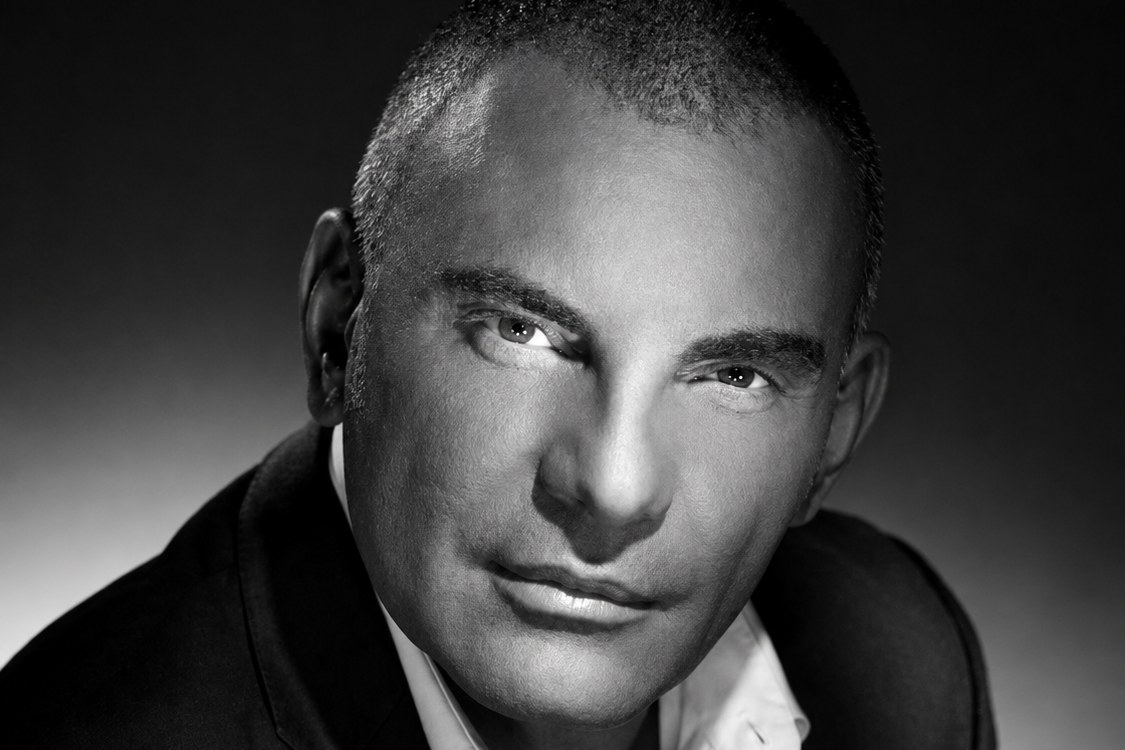 Retrato em preto e branco de um homem de terno escuro e camisa branca aberta, olhando para a câmera.