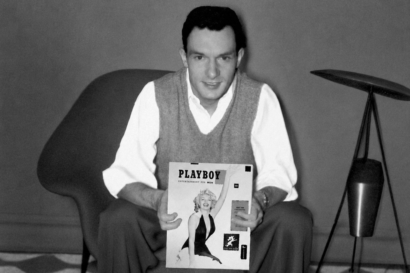 Hugh Hefner jovem, de colete de lã e camisa branca, sentado numa poltrona moderna segurando a primeira edição da Playboy com Marilyn Monroe na capa, ao lado de um abajur de tripé.