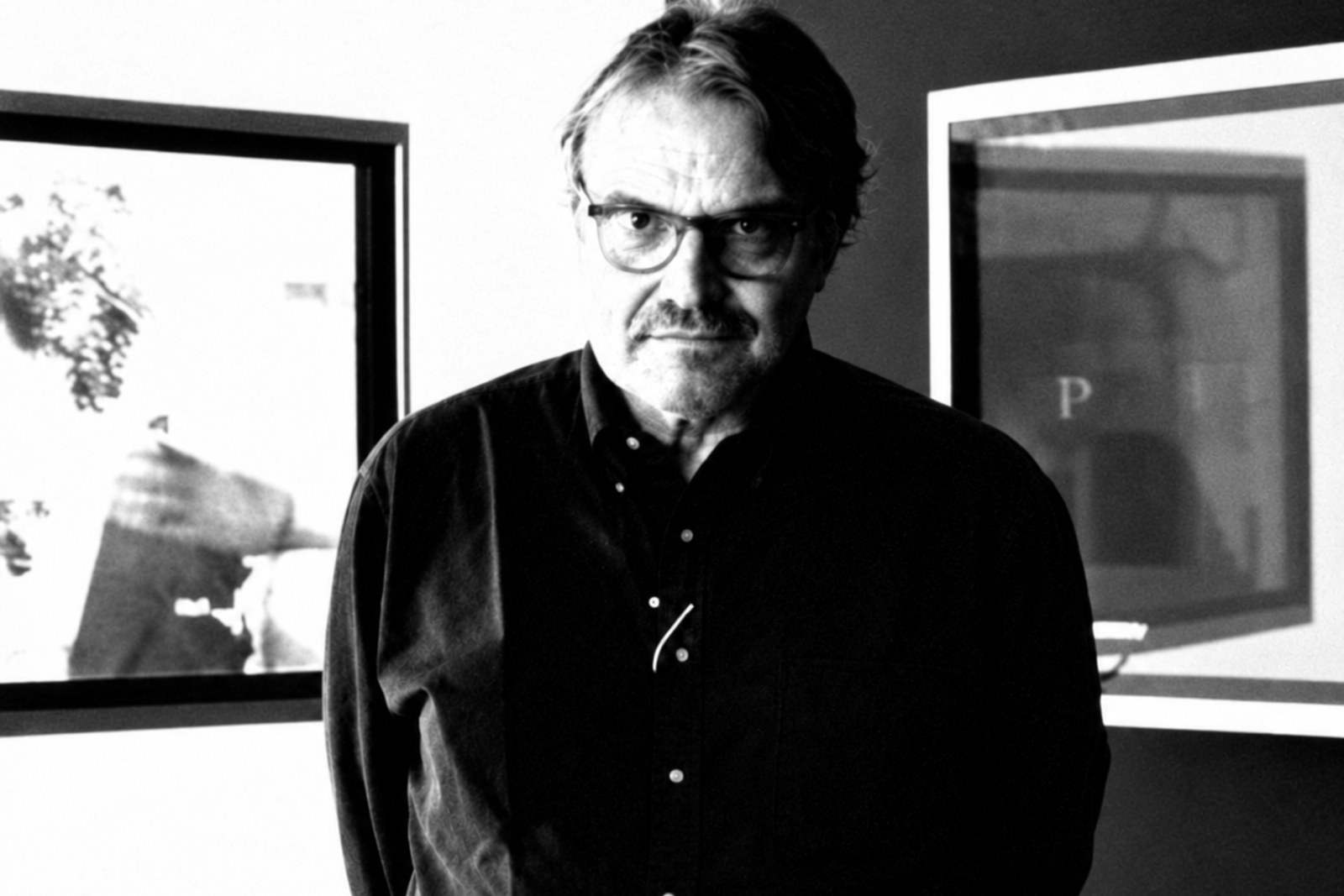 Oliviero Toscani de camisa escura, braços atrás do corpo, diante de duas fotografias emolduradas numa parede dividida entre branco e preto.