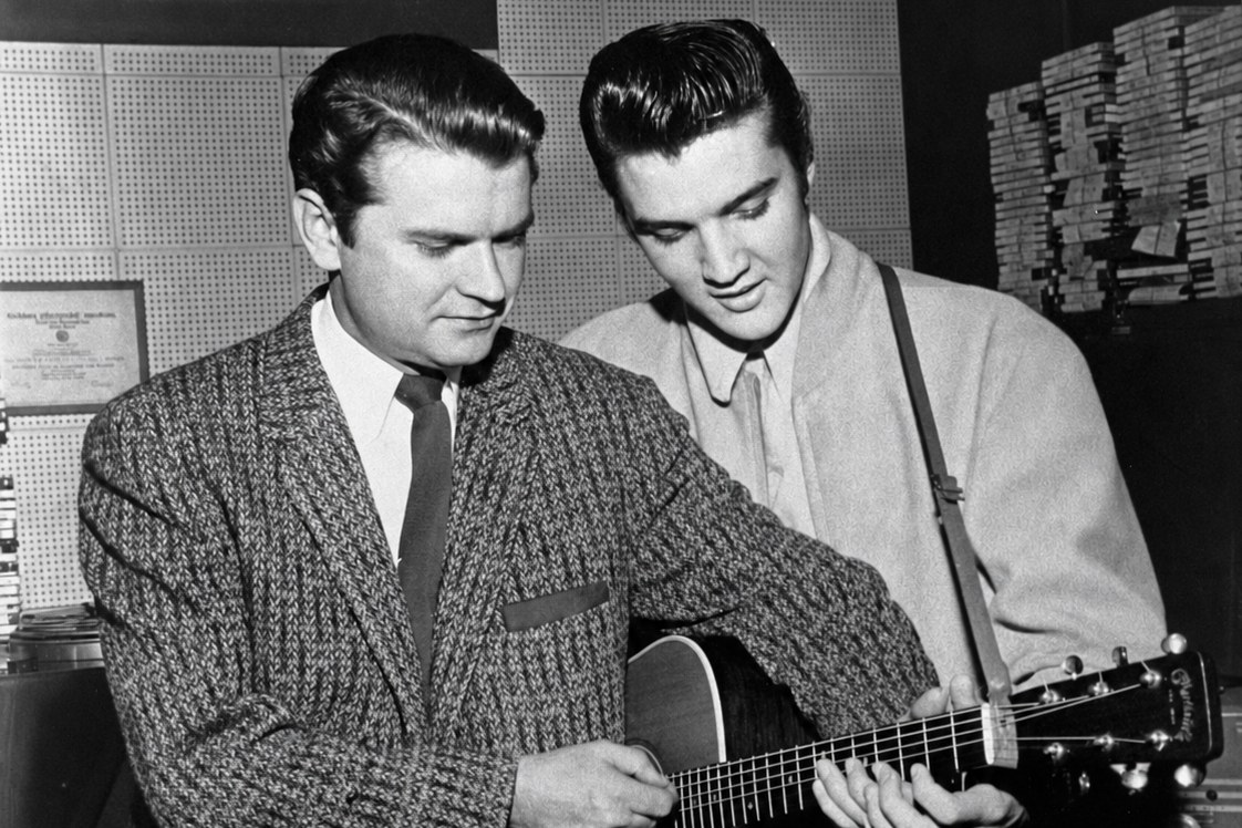 Dois homens de pé num estúdio, um deles segurando o braço de um violão enquanto o outro observa, em imagem monocromática dos anos 1950.
