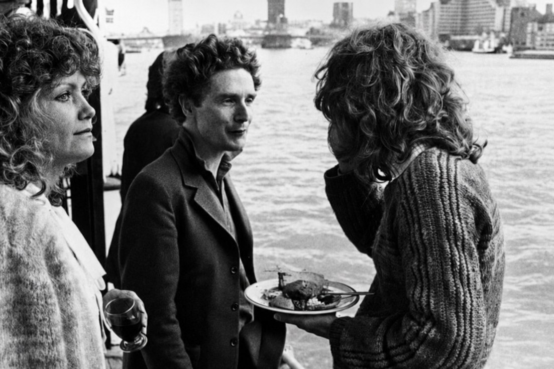 Três pessoas conversando no convés de uma embarcação no Tâmisa, com a margem de Londres ao fundo, em imagem monocromática.